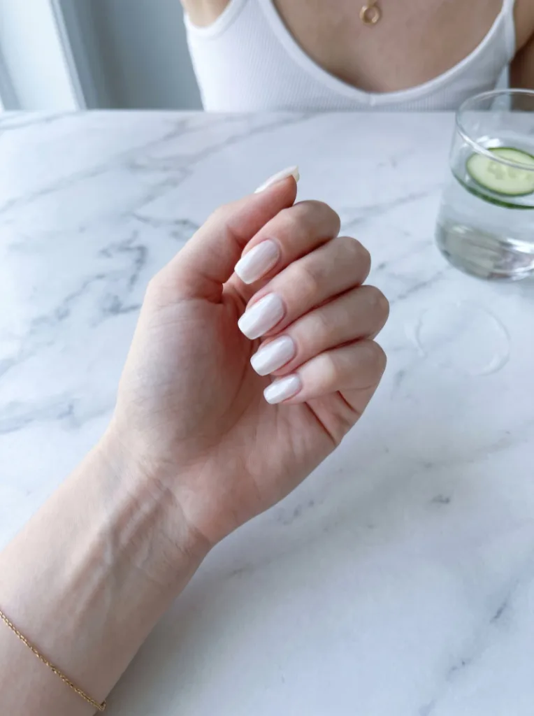 Fair-skinned hand with milky white pearl-finish glazed nail polish resting on white marble beside a glass of water in morning light