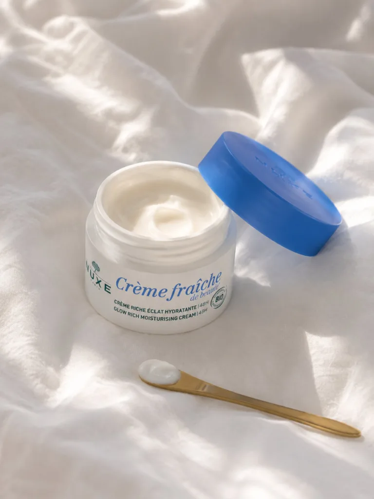 Nuxe Crème Fraîche de Beauté jar open on a rumpled white linen bedsheet with a dab of cream visible