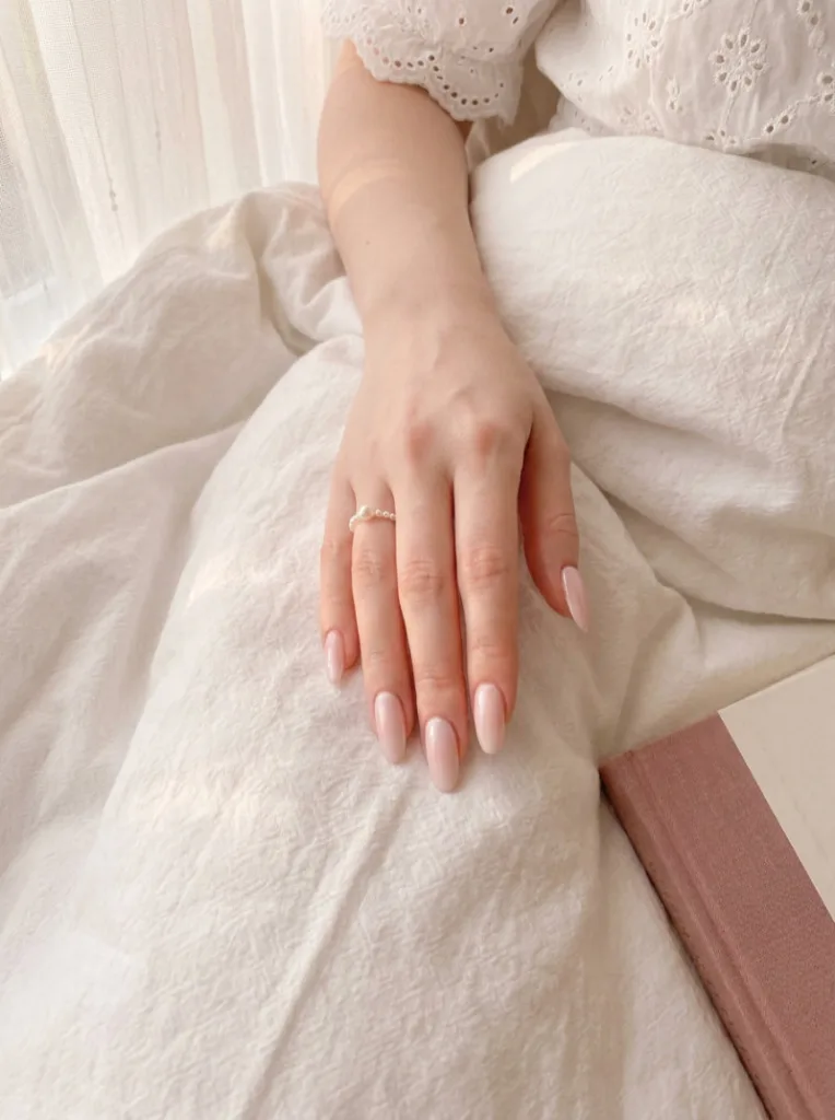 Porcelain-skinned hand with pale blush pink high-gloss nail polish and pearl ring resting on white cotton duvet
