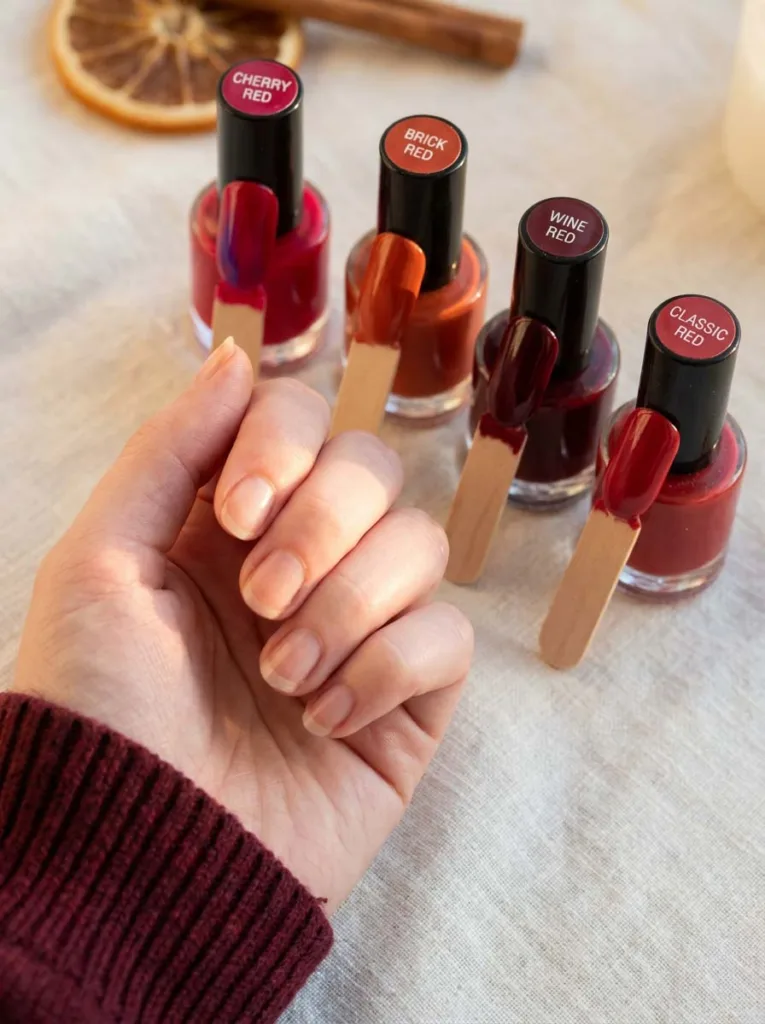 Four red nail polish shades, cherry, brick, crimson, and wine red, beside a manicured hand for skin tone matching