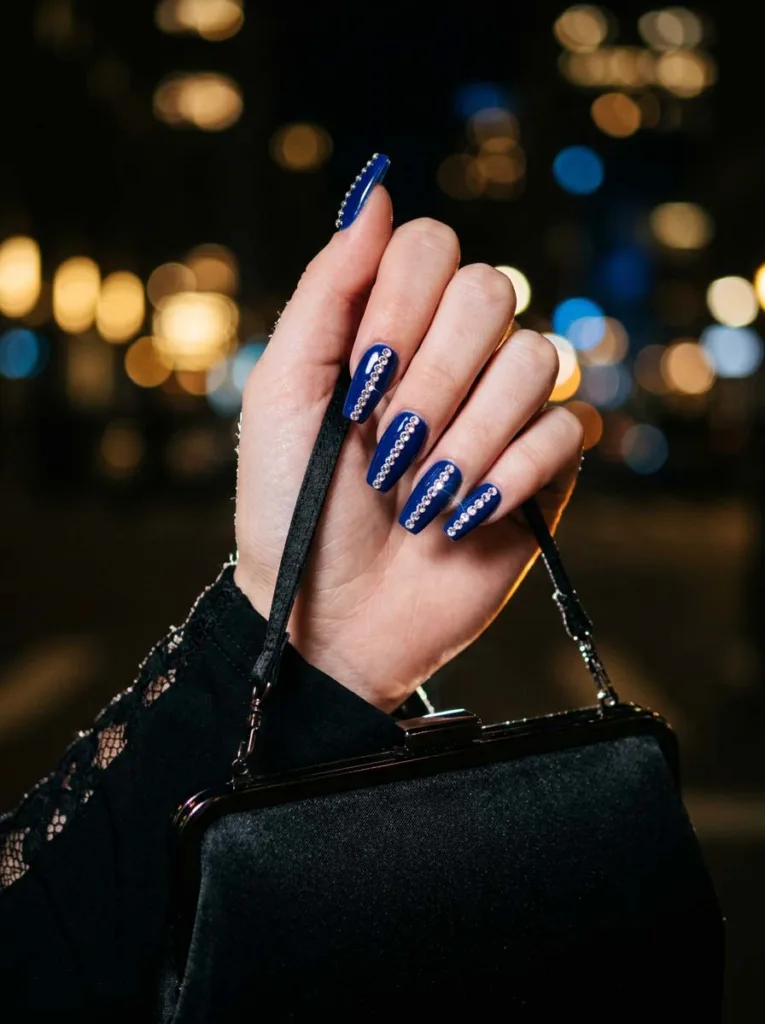 Fair-skinned hand with royal blue rhinestone evening nails holding a black clutch bag