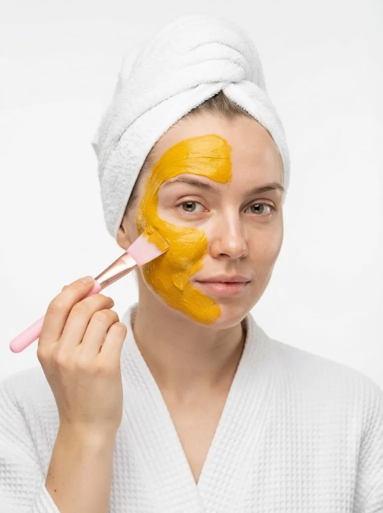 Fair-skinned woman in a white robe applying a vivid golden turmeric face mask to one cheek with a flat silicone brush, looking directly at camera