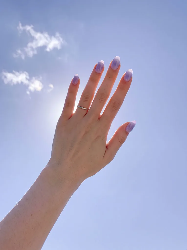 Fair-skinned hand with lavender-to-white aura gradient oval nails reaching upward against a pale blue sky with natural backlight