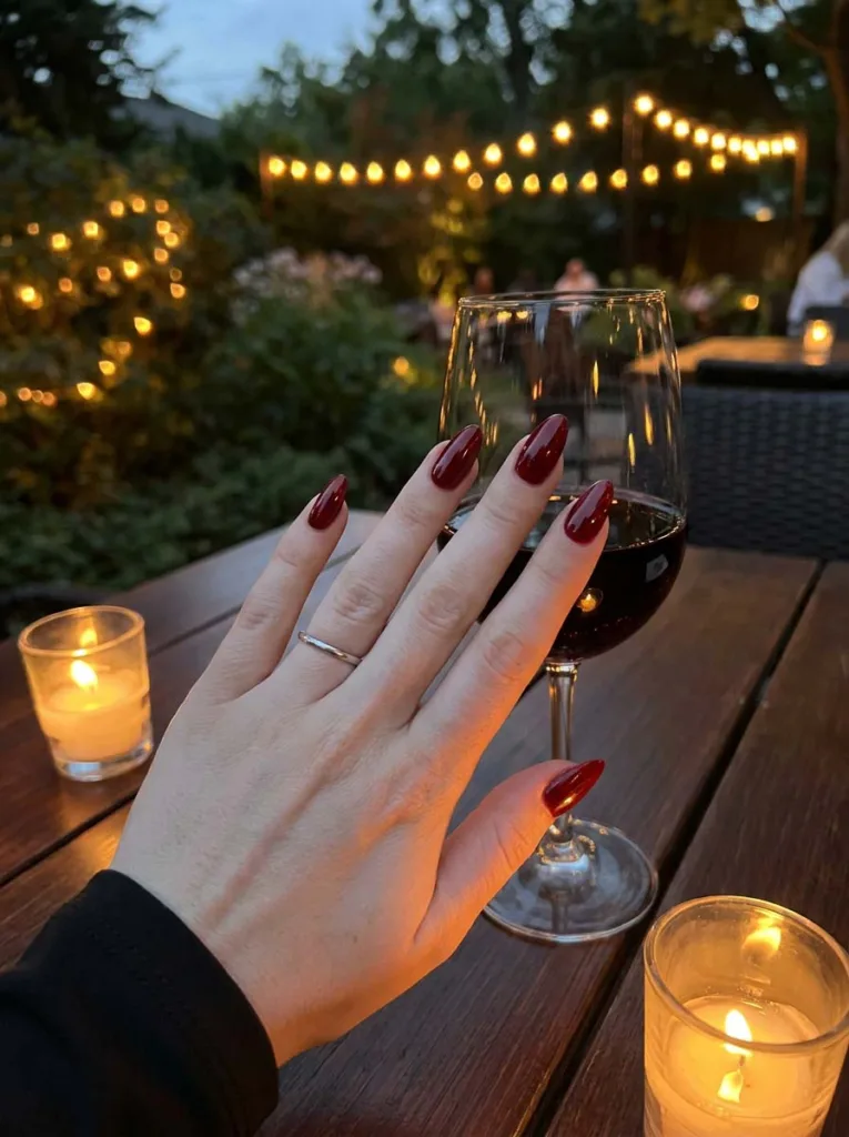 Glossy cherry red summer nails resting near a wine glass at an evening dinner table