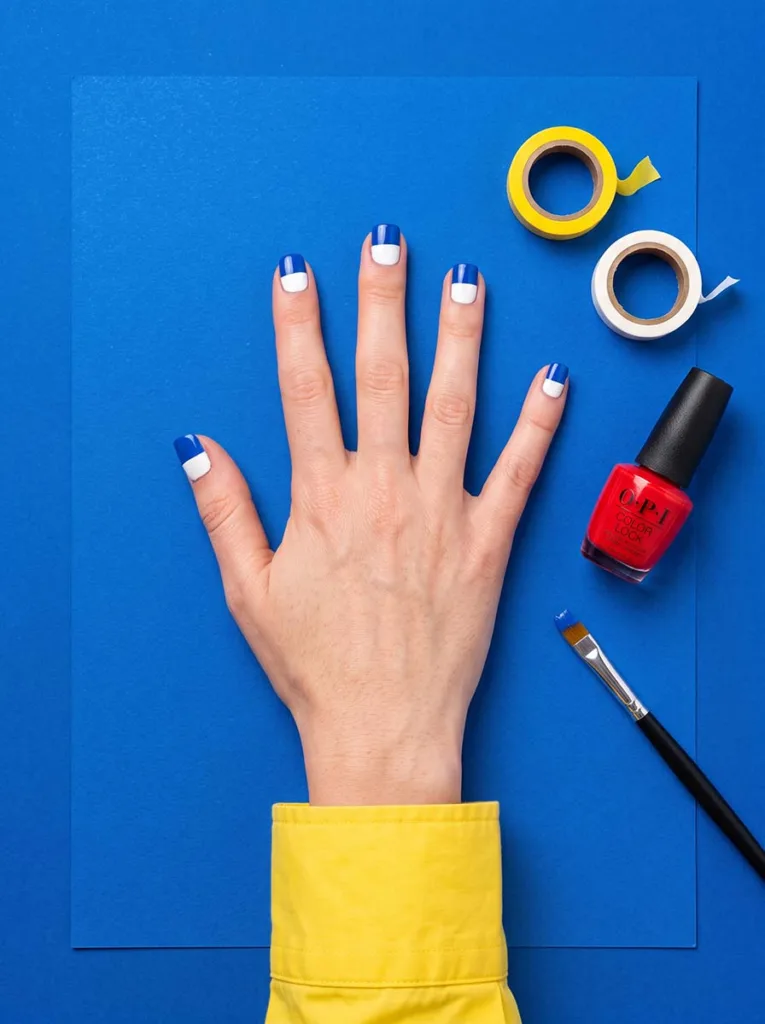 Fair-skinned hand with cobalt blue and white color block short square nails spread flat on bright blue paper with tape and polish beside it