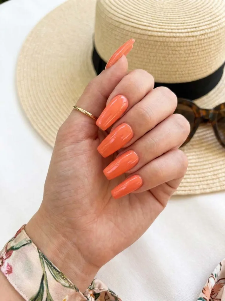 Hand with long coral coffin-shaped orange nails resting on sunglasses by a pool