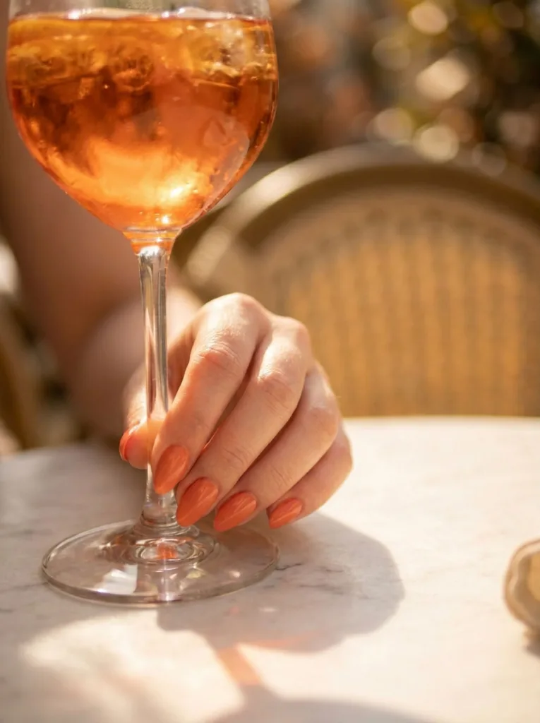 Coral orange summer nails holding an Aperol spritz glass at an outdoor café table