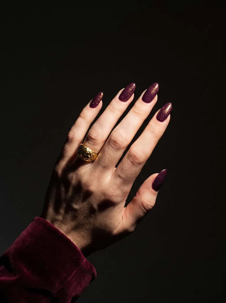 Fair-skinned hand with deep plum berry shimmer almond nails extended toward camera against dark background in dramatic single spotlight
