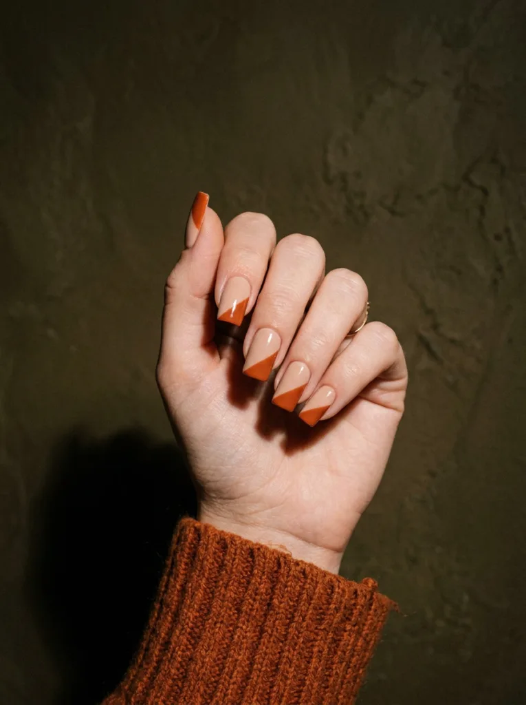 diagonal color block nail art nude and terracotta on square fair skin nails against textured dark wall editorial