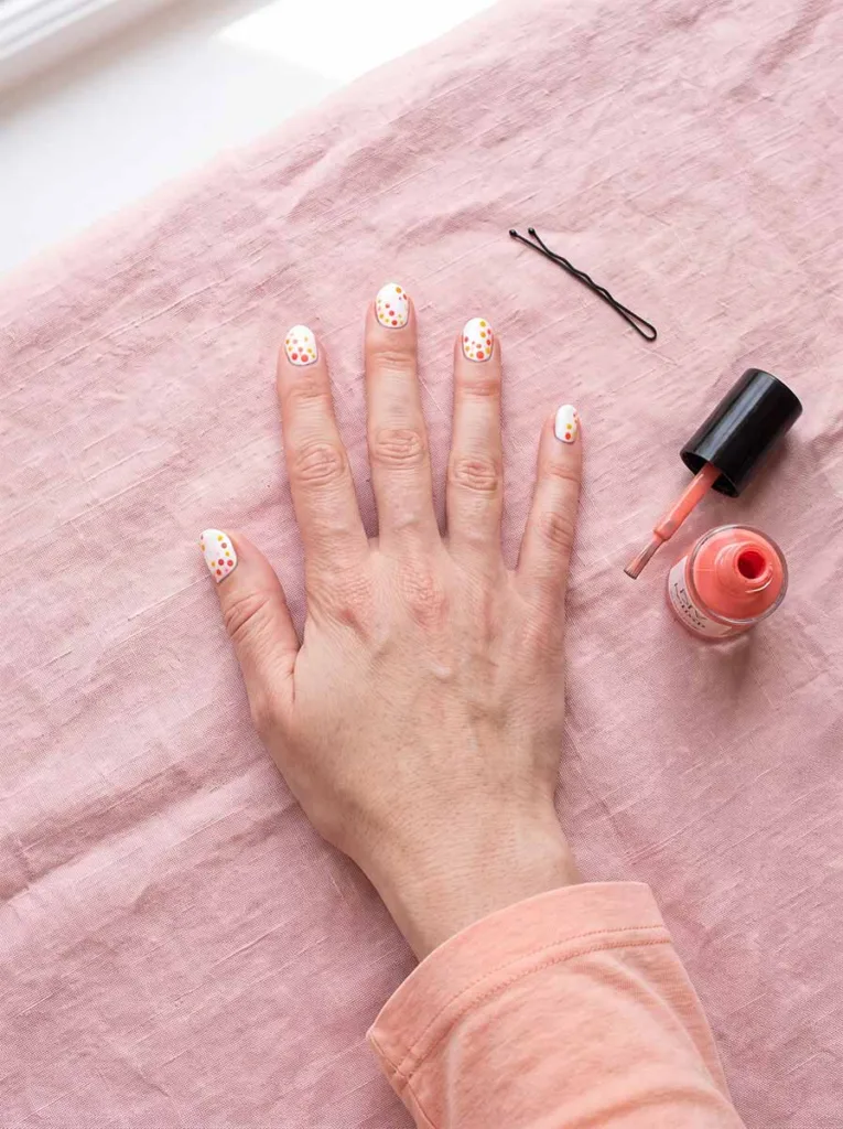 Fair-skinned hand with white base coral and yellow dotticure nails on pink linen with bobby pin and open nail polish bottle beside it