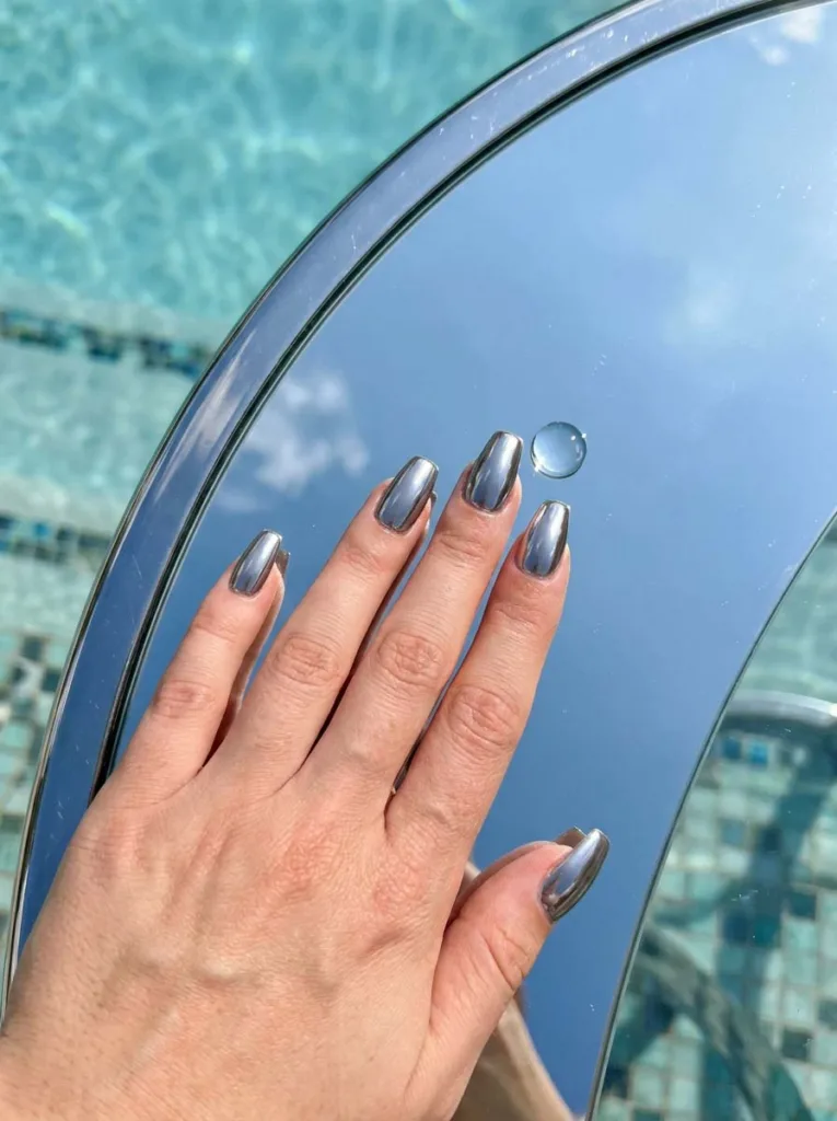 Glazed chrome summer nails reflecting sky and pool water on a glass table