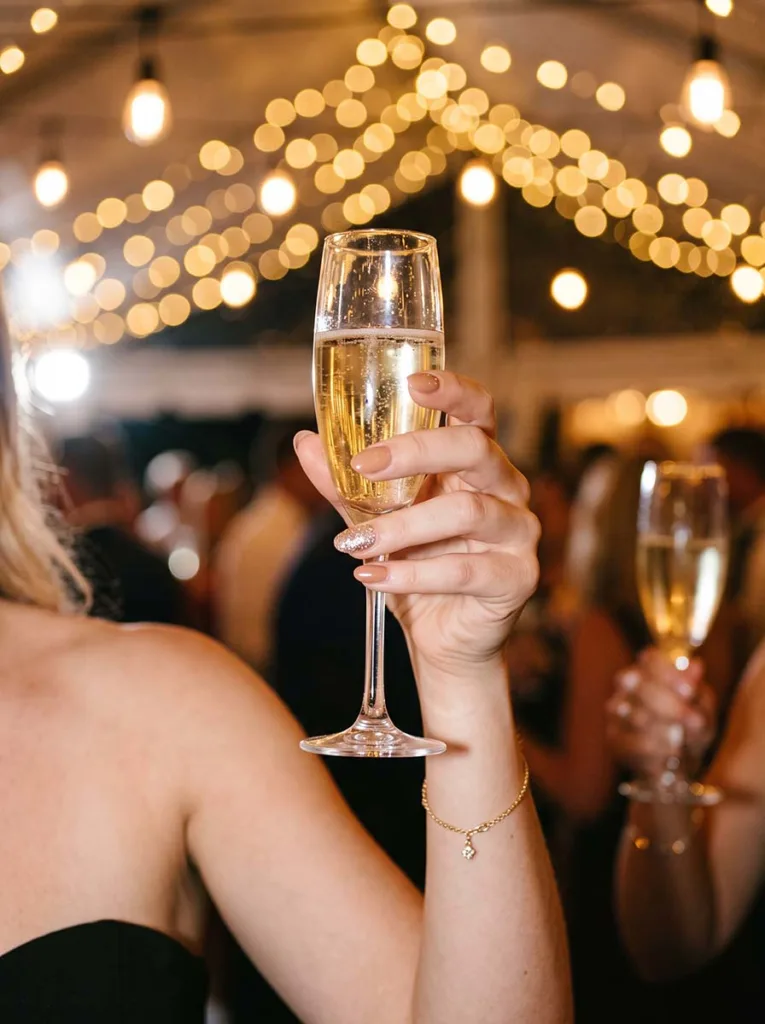Fair-skinned hand with champagne gold glitter accent ring finger nail raising a slim champagne flute in warm string-light party setting