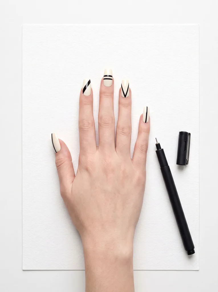 Fair-skinned hand with long square graphic black line art nails spread flat on white watercolor paper beside a technical drawing pen