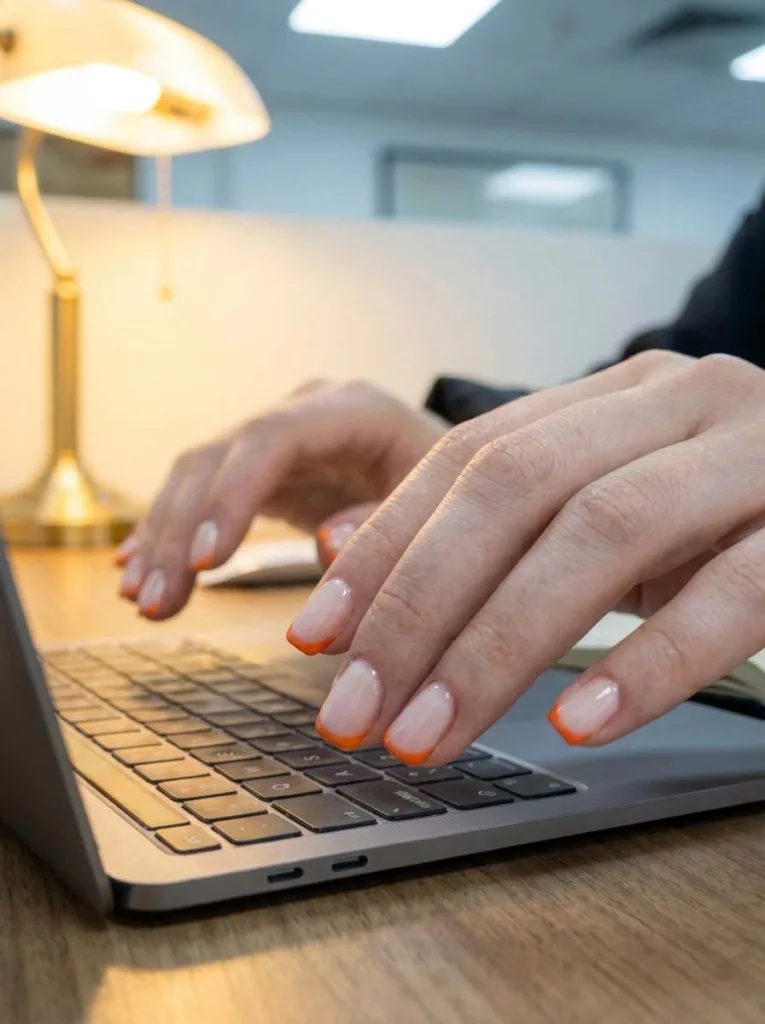 Hand with micro French orange tip nails typing at an office desk