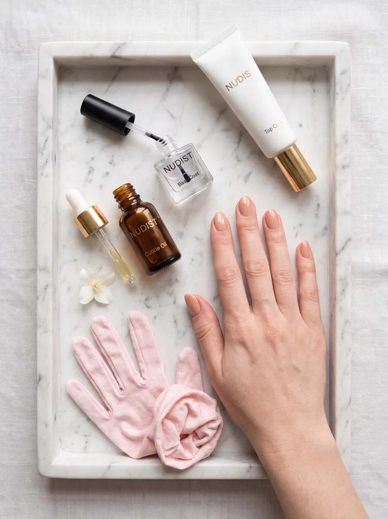Fair-skinned hand with nude manicure resting on white marble tray beside base coat, cuticle oil, top coat, and cotton gloves
