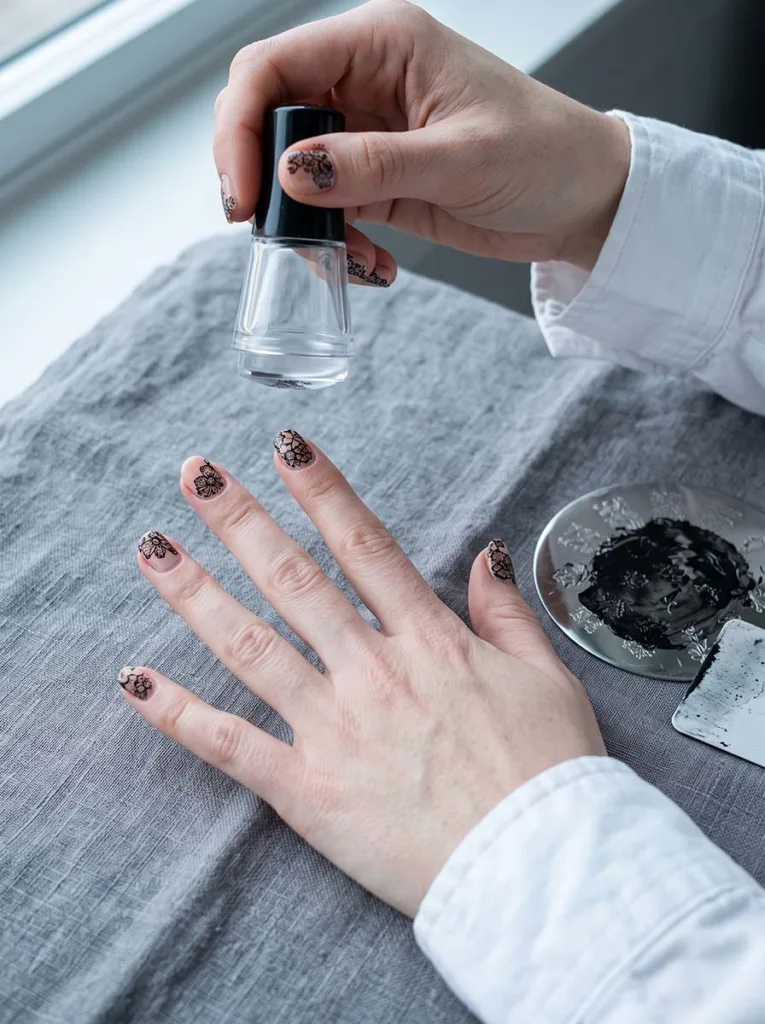Fair-skinned hands showing nail stamping process — completed black floral lace design on nude nails on left hand, stamper tool held in right hand, metal plate visible on grey linen