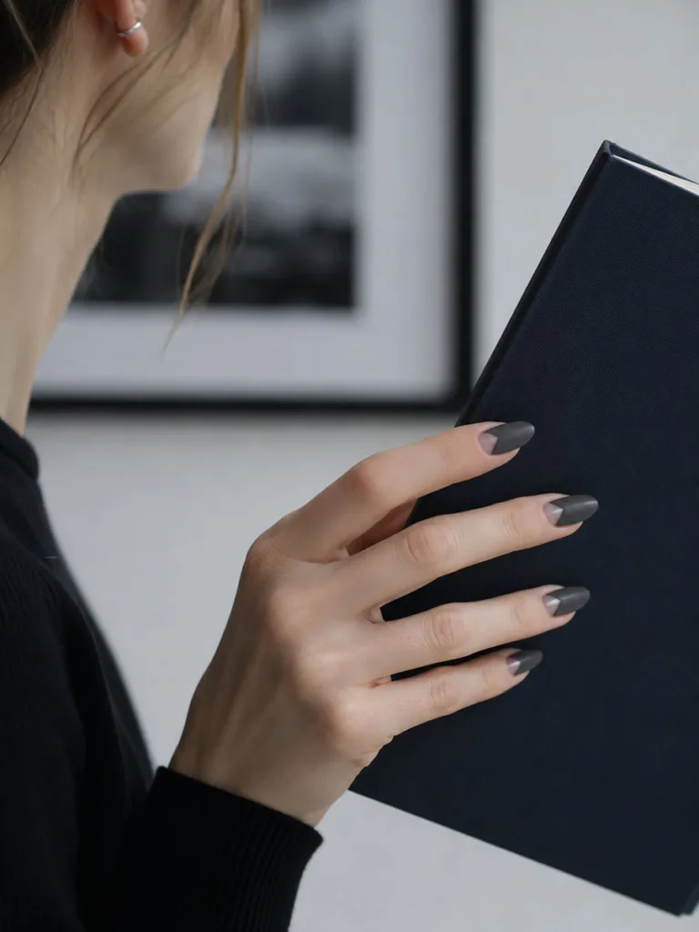 Fair-skinned hand with charcoal negative space almond nails holding a dark navy book, shot from side at low angle