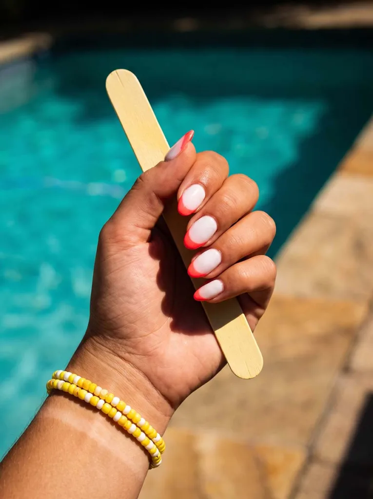 Fair-skinned hand with neon coral French tip nails holding a wooden popsicle stick beside a turquoise pool in direct summer sun
