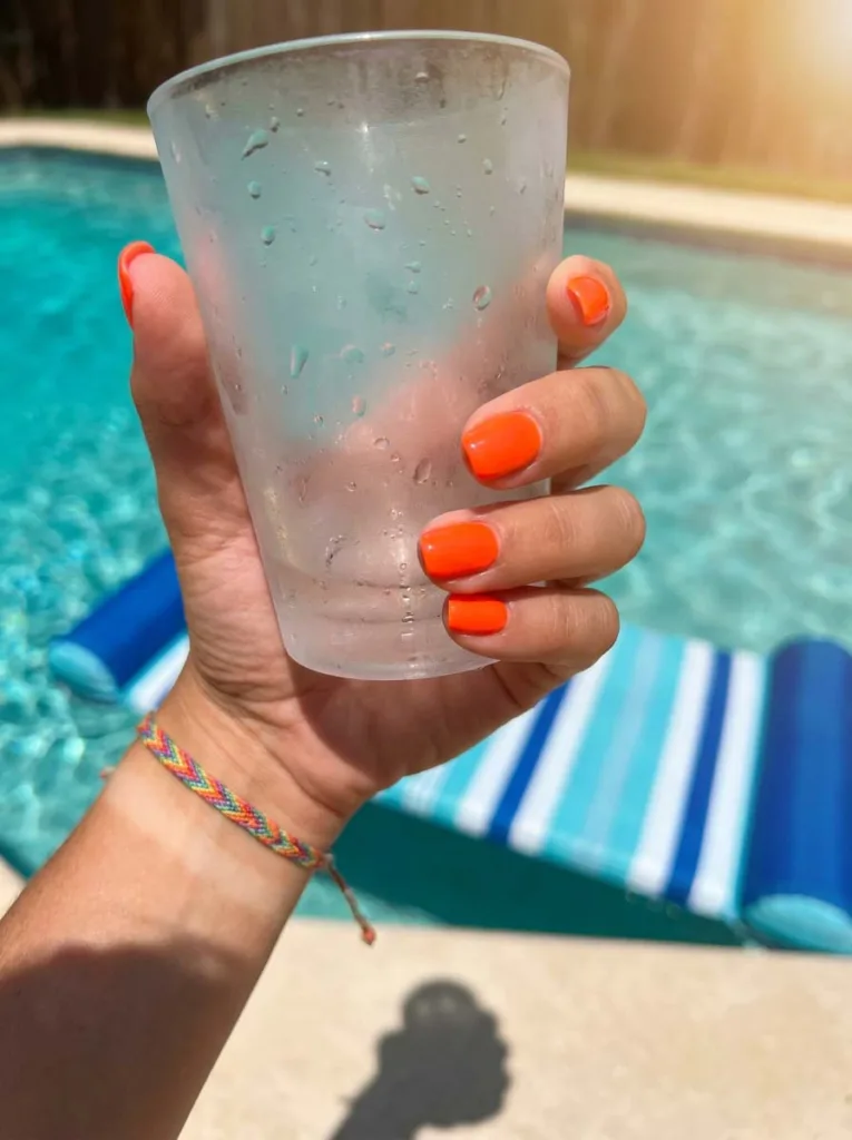 Hand with neon orange nail polish holding a drink by a pool