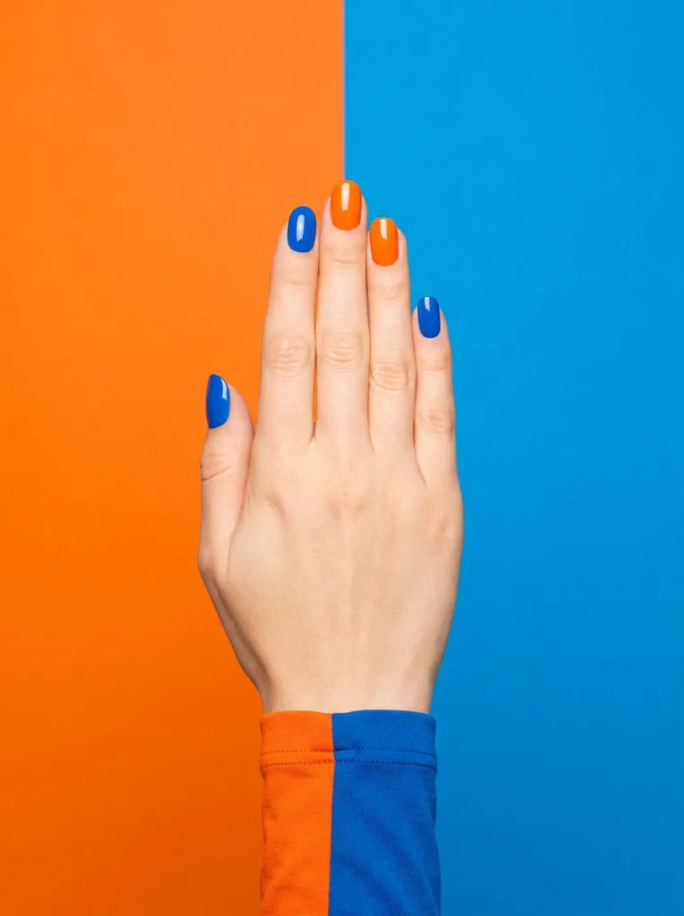 Hand with alternating orange and blue nail polish against a matching color block background
