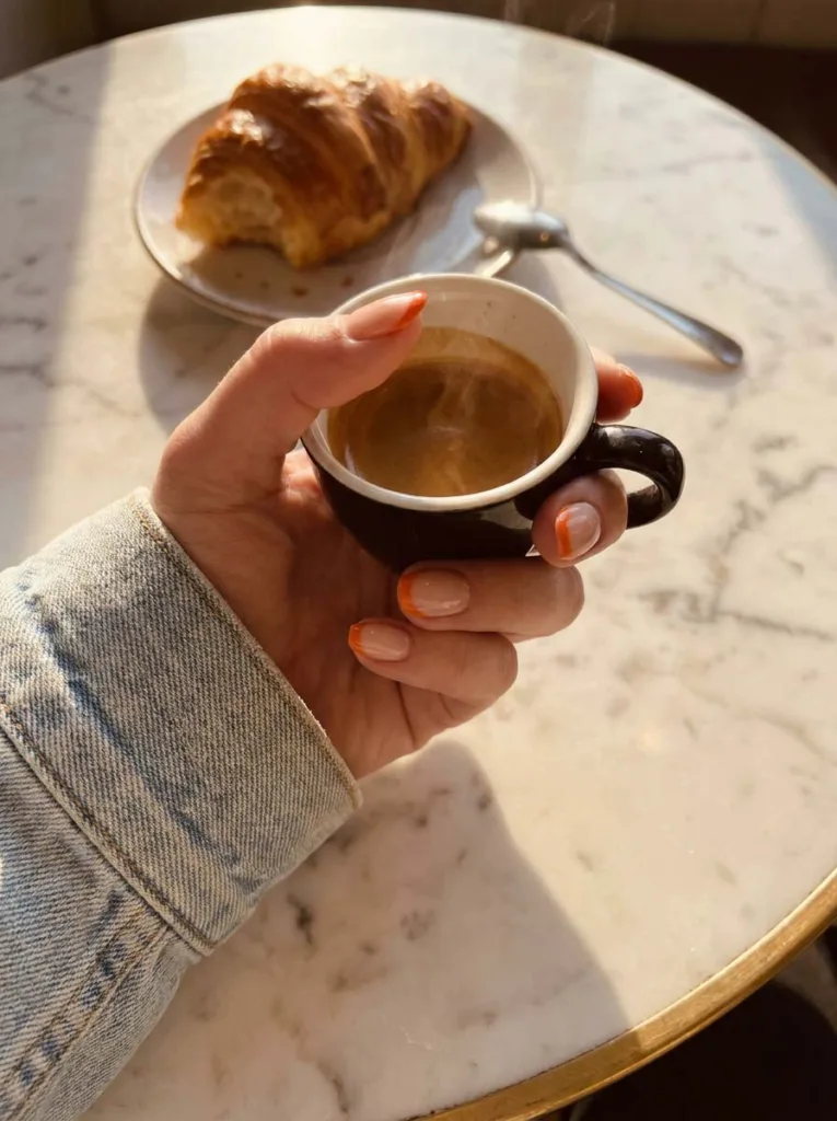 Hand with orange French tip nails holding a coffee cup at a café table