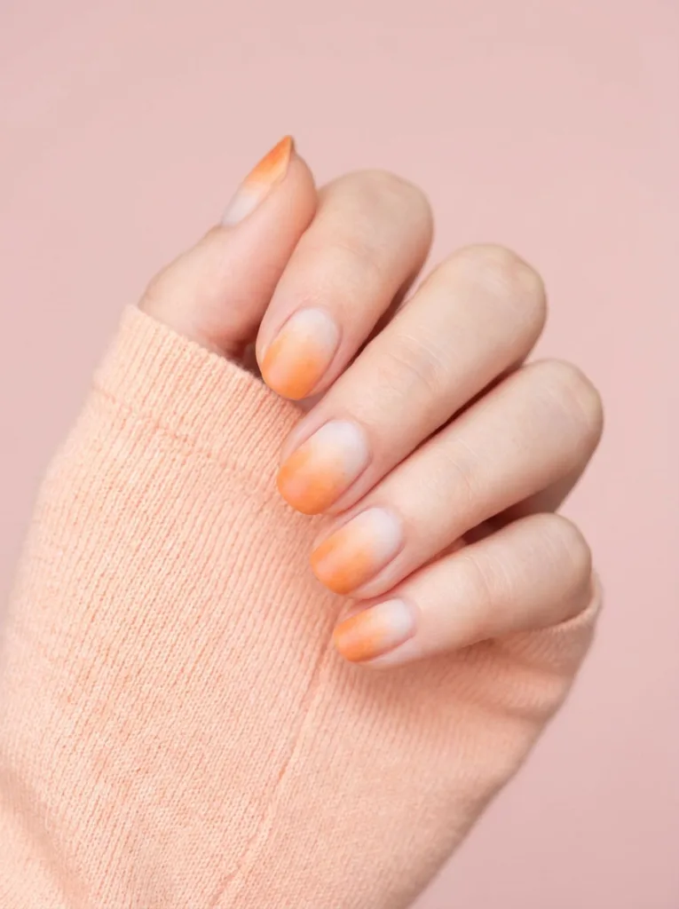 Hand with soft orange ombre nail gradient against a blush pink background