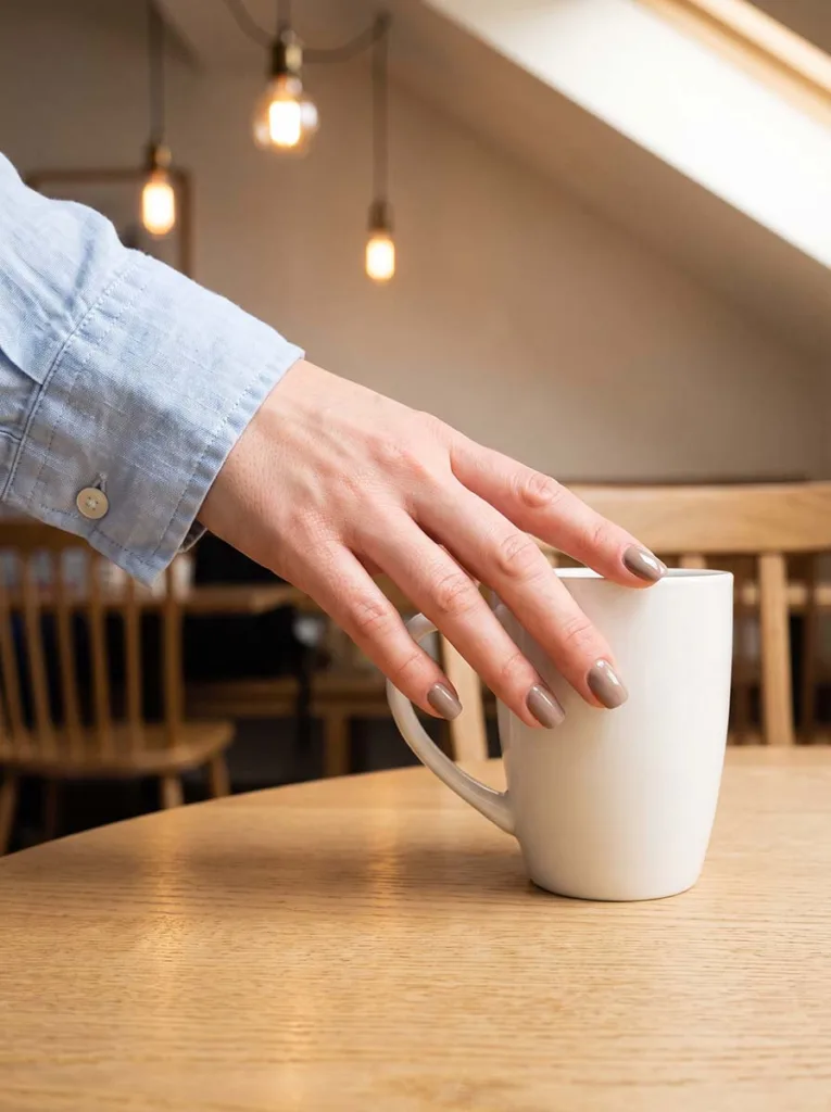 Fair-skinned hand with short greige nails reaching for a white ceramic coffee mug on a light oak café table