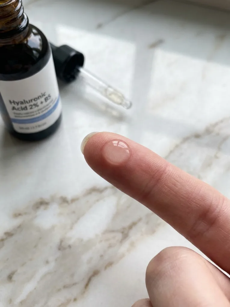 The Ordinary hyaluronic acid serum dropper bottle with a single clear drop balanced on a fair-skinned fingertip on white marble