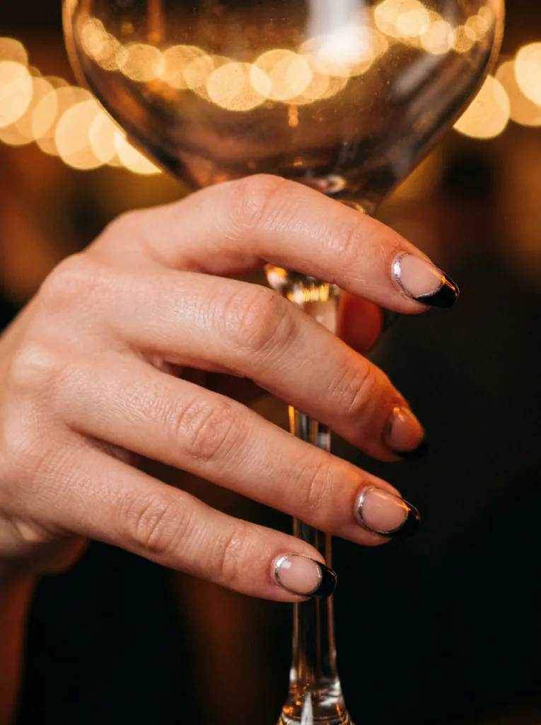 Close-up of a hand holding a cocktail glass stem, black French tips with silver lining