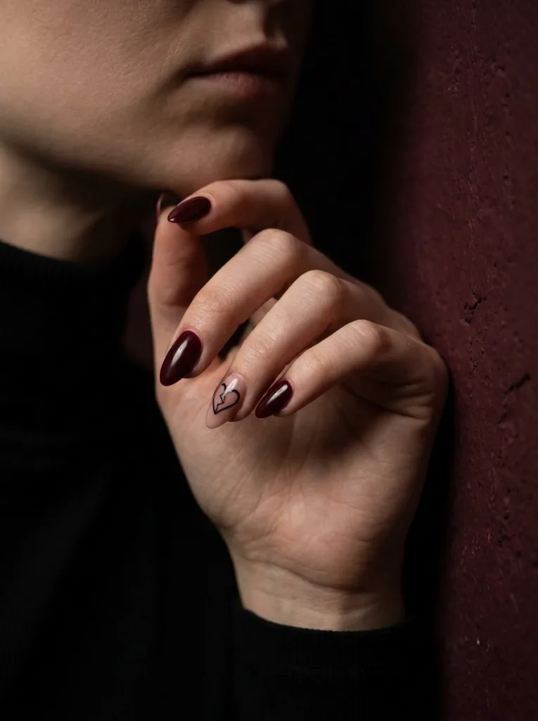 Fair-skinned hand with burgundy almond nails and broken heart nail art accent