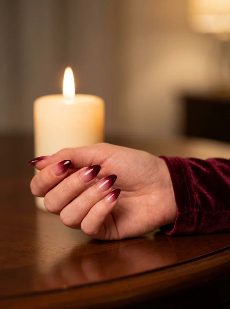 Fair-skinned hand with burgundy ombre almond nails near a lit candle