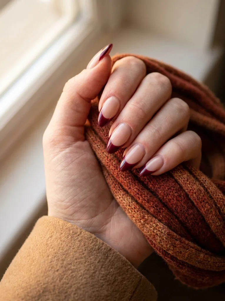 Glossy burgundy French tips on a nude base, hand resting on a warm rust-colored knit scarf