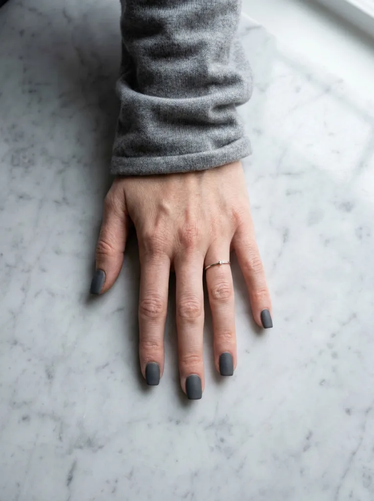 Fair-skinned hand with matte charcoal grey square winter nails resting on a marble table