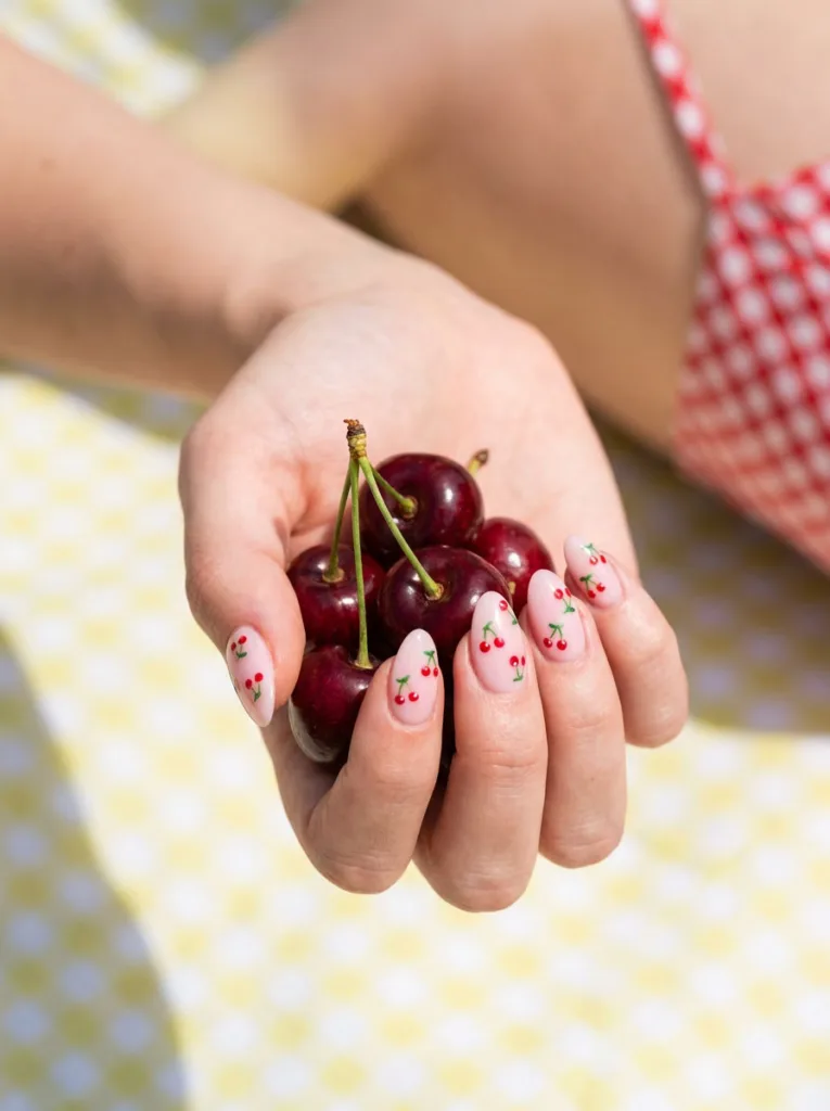 Fair-skinned hand with pink almond nails and hand-painted cherry nail art holding cherries