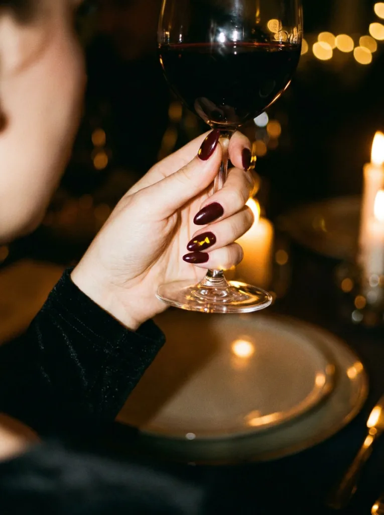 Fair-skinned hand holding a wine glass with deep burgundy simple winter nails at a candlelit table
