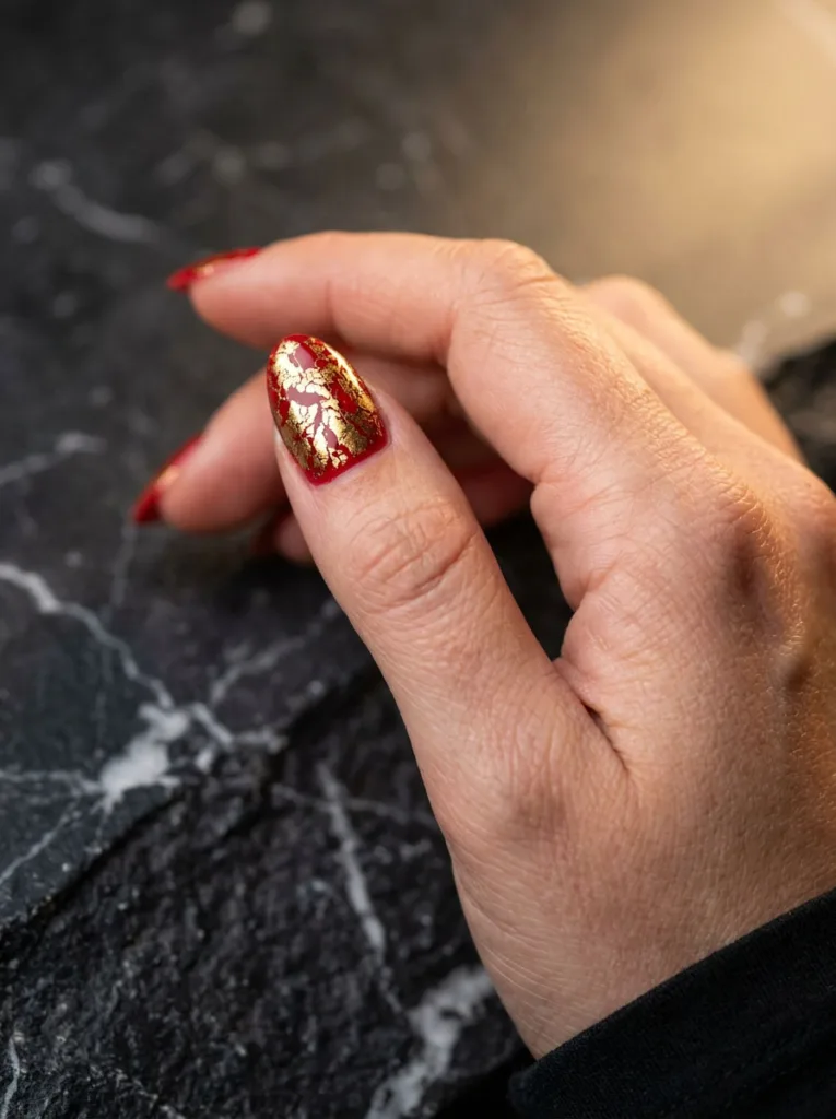 Fair-skinned hand with red almond nails and cracked gold foil accent nail on marble