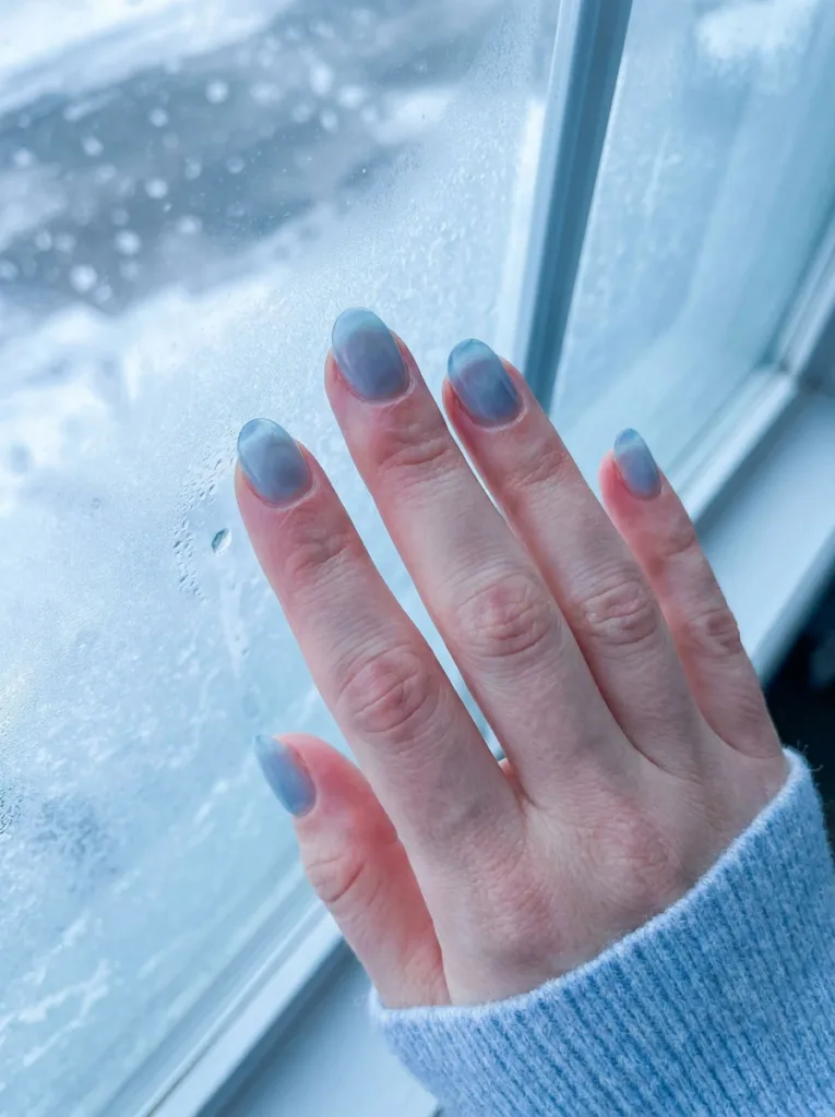 Fair-skinned hand with icy blue sheer simple winter nails near a frosted winter window