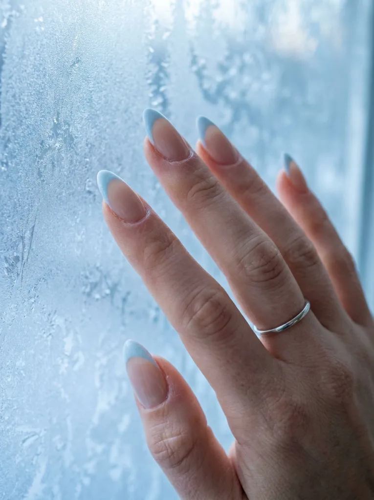 Pale frost blue French tips on a sheer nude base near a frosted winter window