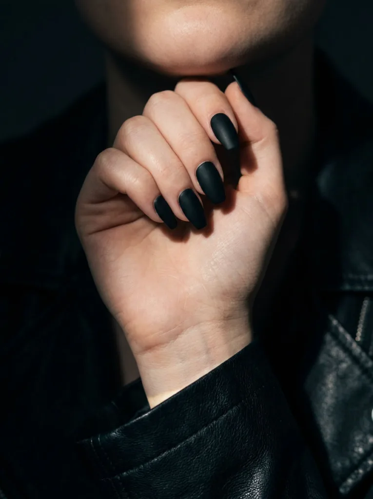 Fair-skinned hand near chin showing matte black simple winter nails against a dark backdrop