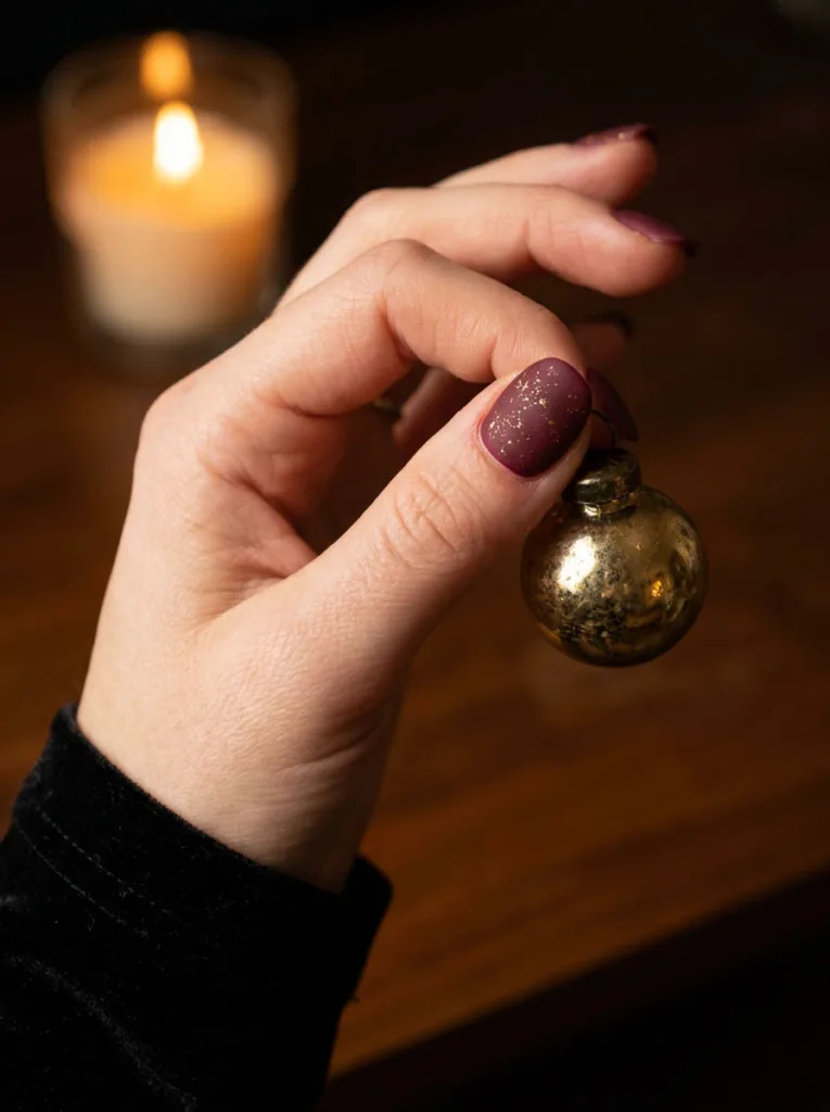 Fair-skinned hand with matte burgundy snowflake christmas nails holding a gold glass ornament.