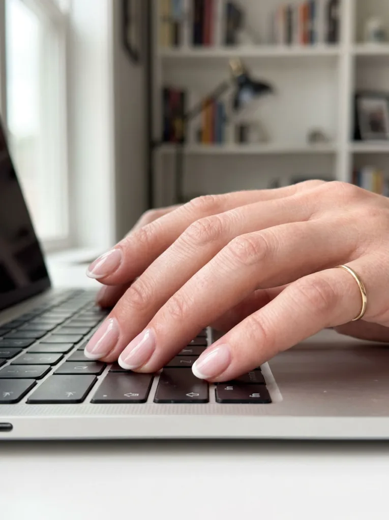 Thin white micro French tips on short natural nails, hand resting on a laptop keyboard