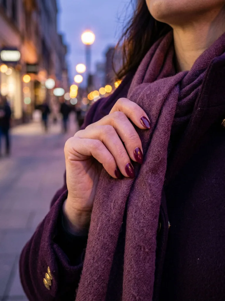 Fair-skinned hand adjusting a scarf showing plum wine simple winter nails at dusk