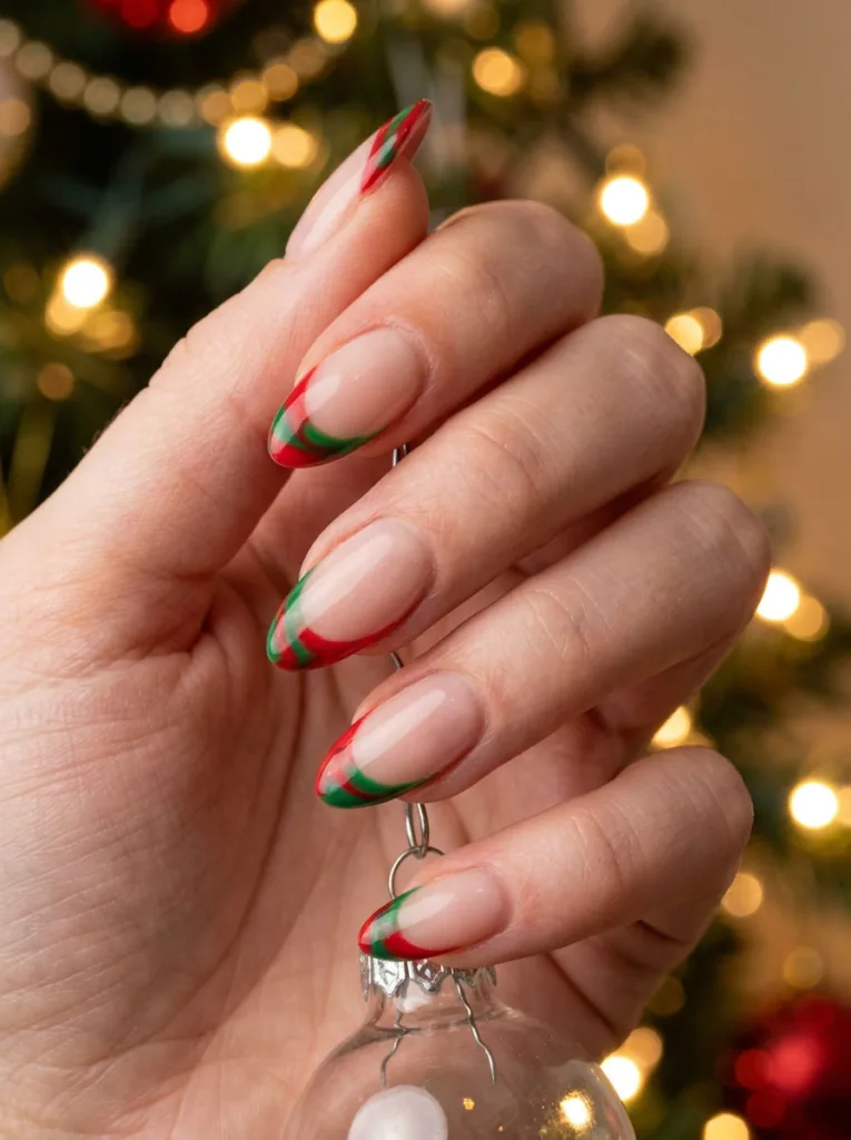 Red and green swirled French tips on winter nails, hand holding a Christmas ornament near a lit tree