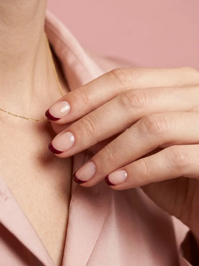 Hand with a burgundy half-moon manicure adjusting a gold necklace