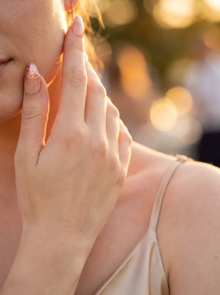 Fair-skinned hand with rose gold glitter almond nails touching cheek at golden hour