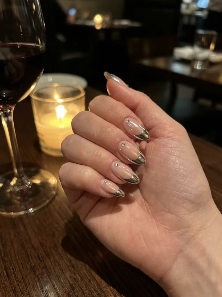 High-shine silver chrome French tips with tiny rhinestones, hand resting beside candlelight at a dinner table