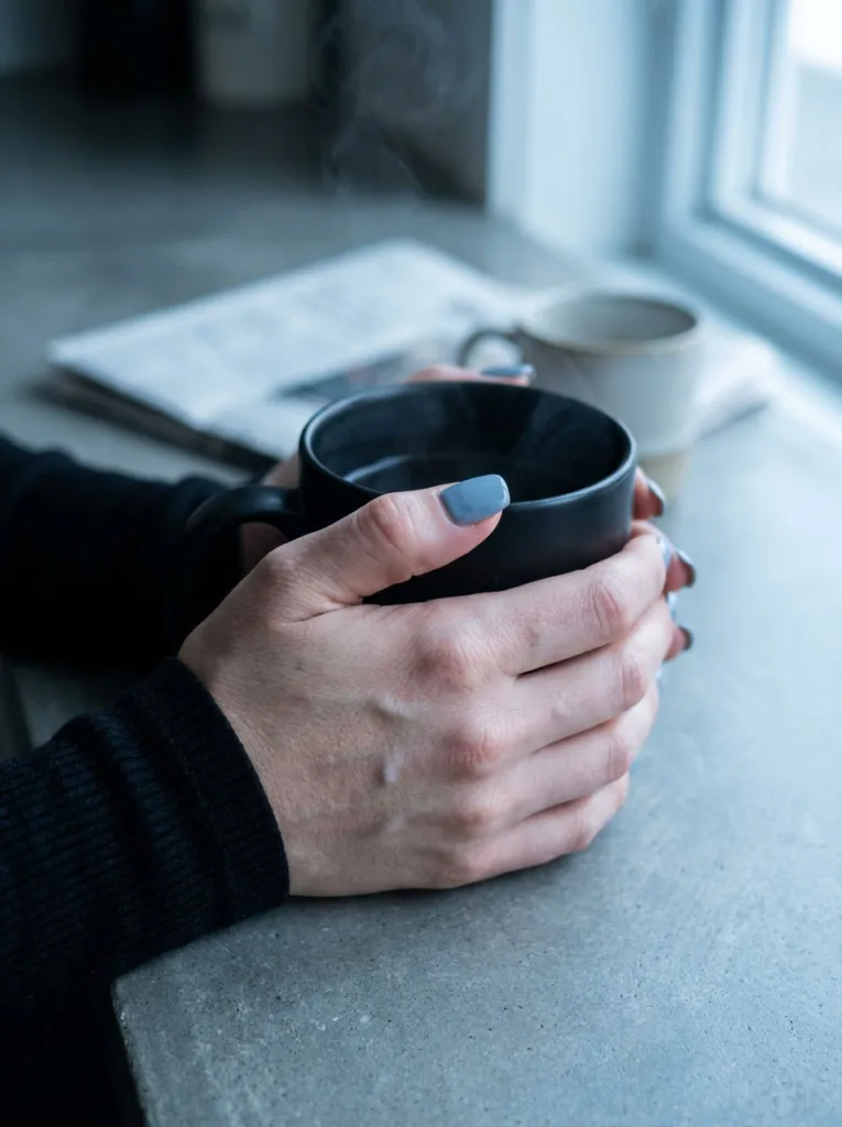 Fair-skinned hand wrapped around a matte black mug showing soft gray simple winter nails