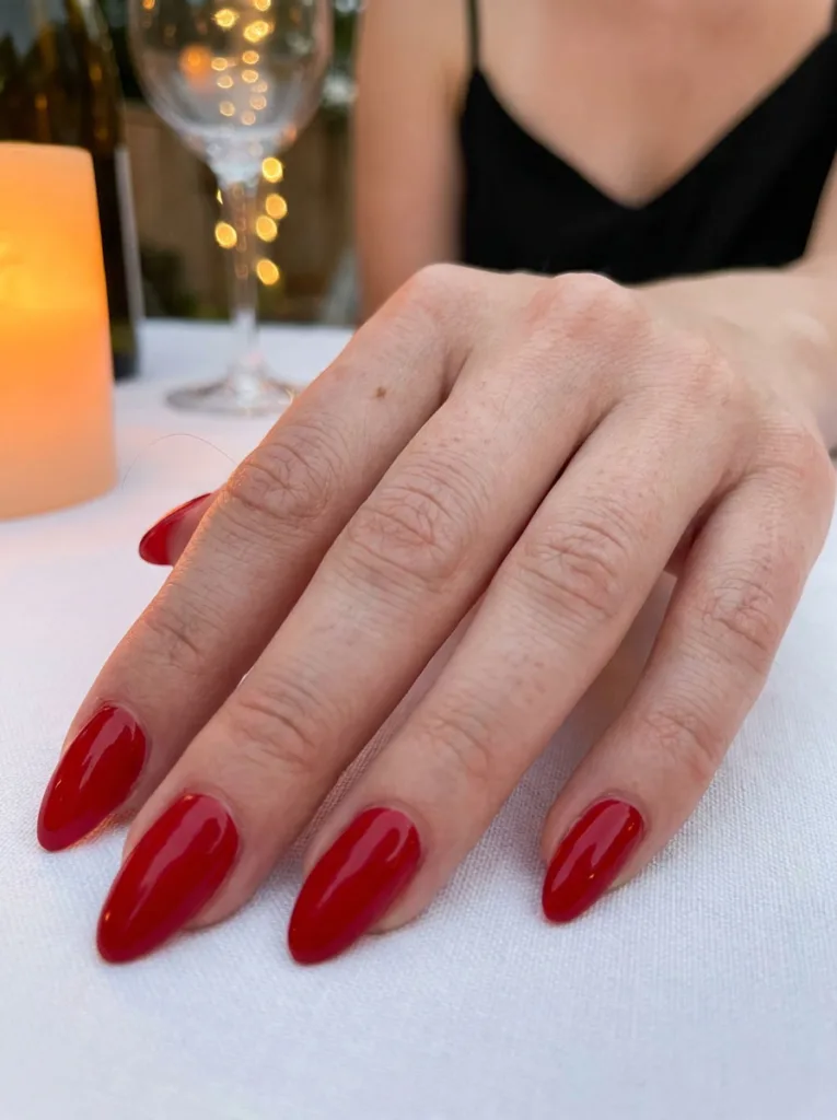 Woman's hand with a classic glossy red manicure resting on a white tablecloth beside a wine glass.