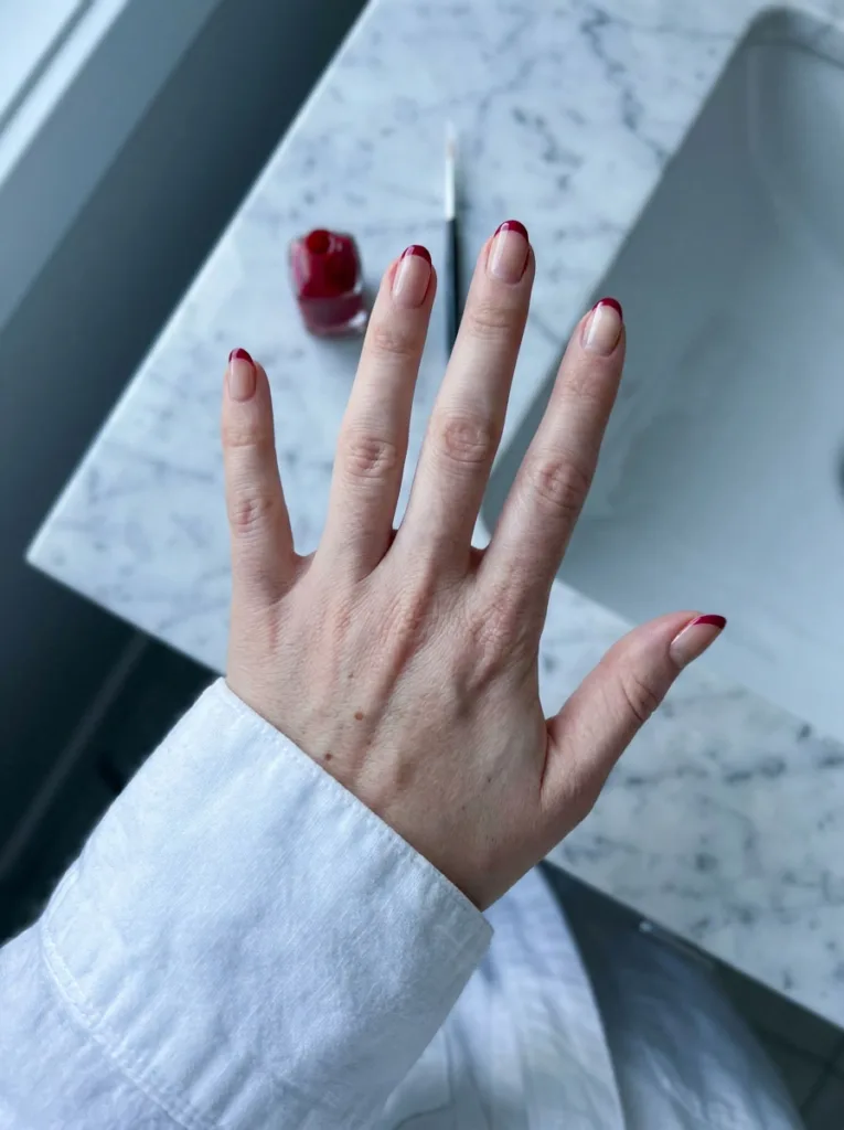 Hand with red French tip nails resting on a bathroom sink beside a nail polish bottle.