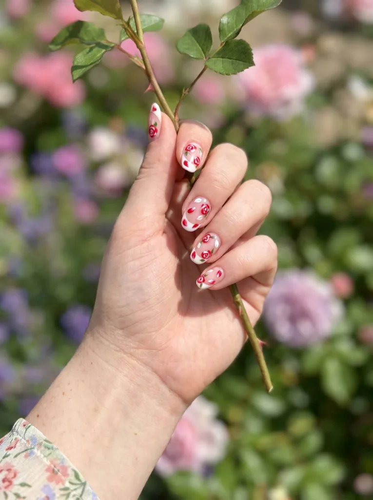 Hand with red floral nail art gently holding a rose stem in a garden.